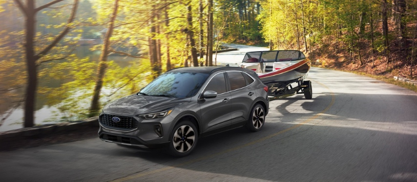  2025 Ford Escape towing a boat on a forested road near a lake.