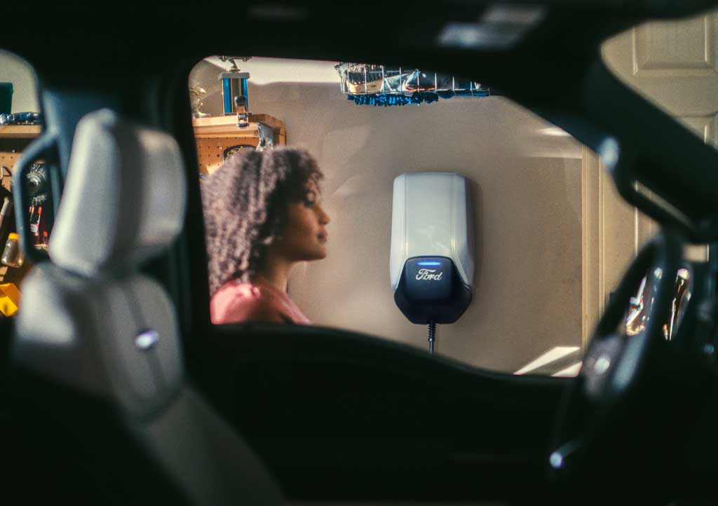 A Ford Connected Charge Station mounted on a garage wall with a woman seen through a Mustang Mach-e window