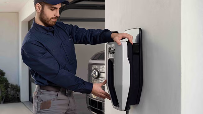 Technician adjusts a Ford-branded wall-mounted EV charger in a garage with a Ford electric vehicle parked behind him.