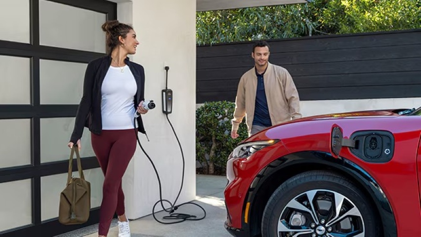 A woman prepares to plug in her red Ford electric vehicle using a wall-mounted home charger as a man approaches with a smile.