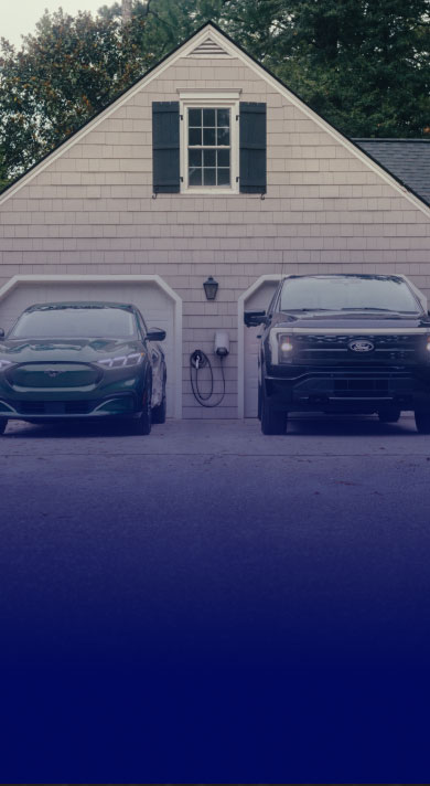 A Mustang Mach-E® and F-150 Lightning® sitting in a driveway with Ford Connected Charge Station hanging on the wall of the garage.
